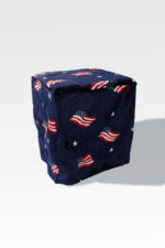 An object resembling an EZ Fold Cremation Stand, cube-shaped and covered in dark blue fabric adorned with U.S. flags and white stars, casts a shadow on a light gray background.