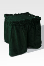 A dark green, square plush ottoman with a ruffled skirt is elegantly set against a white backdrop, evoking timeless charm while resting atop a Stainless Steel Cremation Stand.
