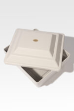 A Cremains Urn Vault, square and white with a ceramic finish, featuring a slightly ajar matching lid, evokes the elegance of a delicate urn against a plain background.