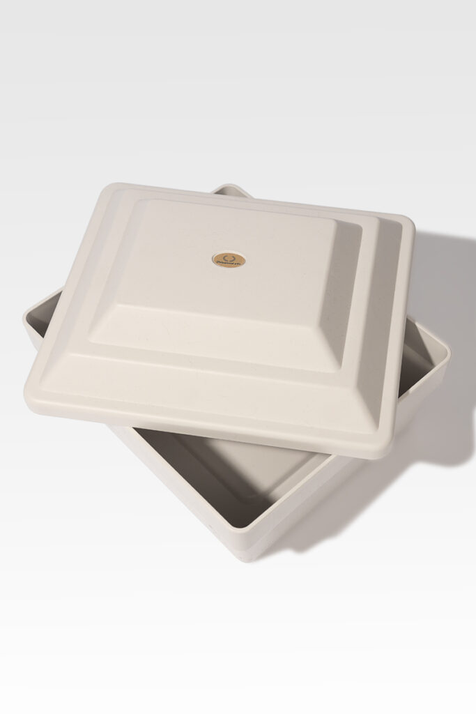 A Cremains Urn Vault, square and white with a ceramic finish, featuring a slightly ajar matching lid, evokes the elegance of a delicate urn against a plain background.