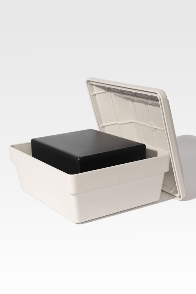 A black cube is positioned inside an open Cremains Urn Vault, which is made of white plastic, set against a light gray background.