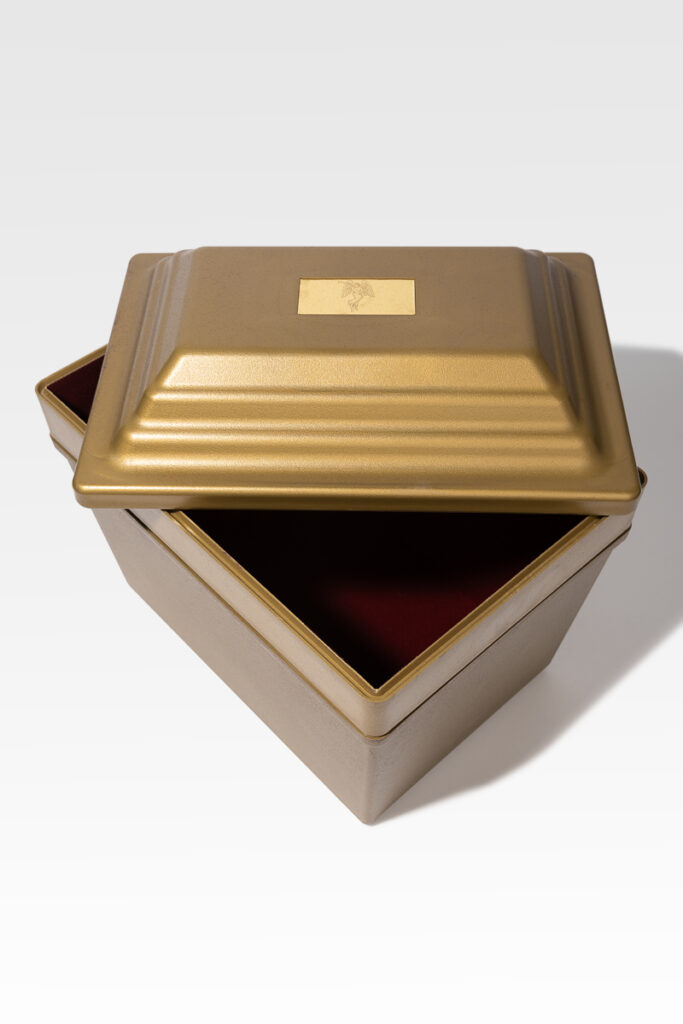 This gold metal box is a Fortress® Urn Vault, featuring a hinged lid, a dark interior lining, and a rectangular blank label on top.