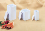 Three white rectangular Infant, Child, and Large Child Temporary Urns with detached lids are displayed in a row on a beige cloth, alongside a small bouquet of assorted flowers in the lower left corner.