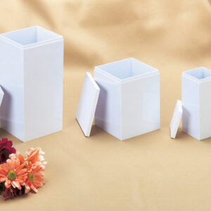 Three white rectangular Infant, Child, and Large Child Temporary Urns with detached lids are displayed in a row on a beige cloth, alongside a small bouquet of assorted flowers in the lower left corner.