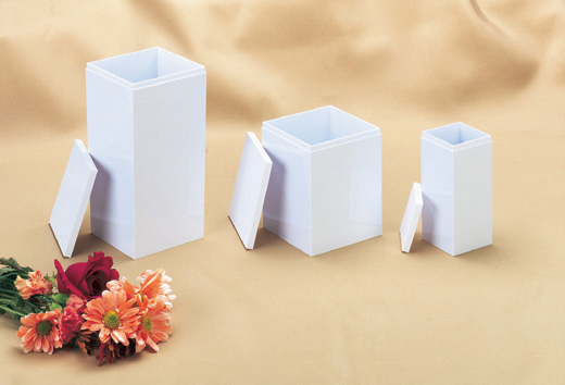 Three white rectangular Infant, Child, and Large Child Temporary Urns with detached lids are displayed in a row on a beige cloth, alongside a small bouquet of assorted flowers in the lower left corner.