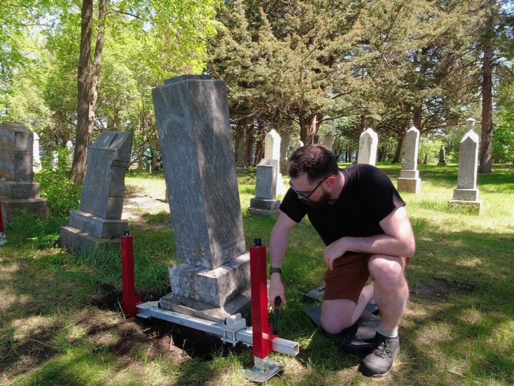 A man adjusts leveling equipment on a tilted gravestone using a Tombstone Jack in a cemetery surrounded by trees and other headstones on a sunny day.
