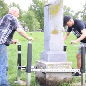 Two men are working to level a tilted gravestone using a Tombstone Jack and other mechanical tools in a grassy cemetery.