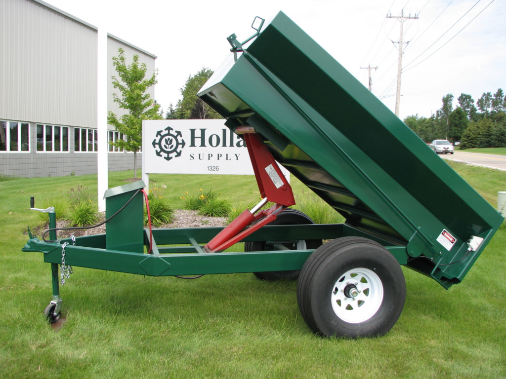 Green utility trailer with hydraulic lift, slightly elevated on grassy terrain, could be among the top 10 products you must have at your cemetery. A white building and company sign stand proudly in the background.