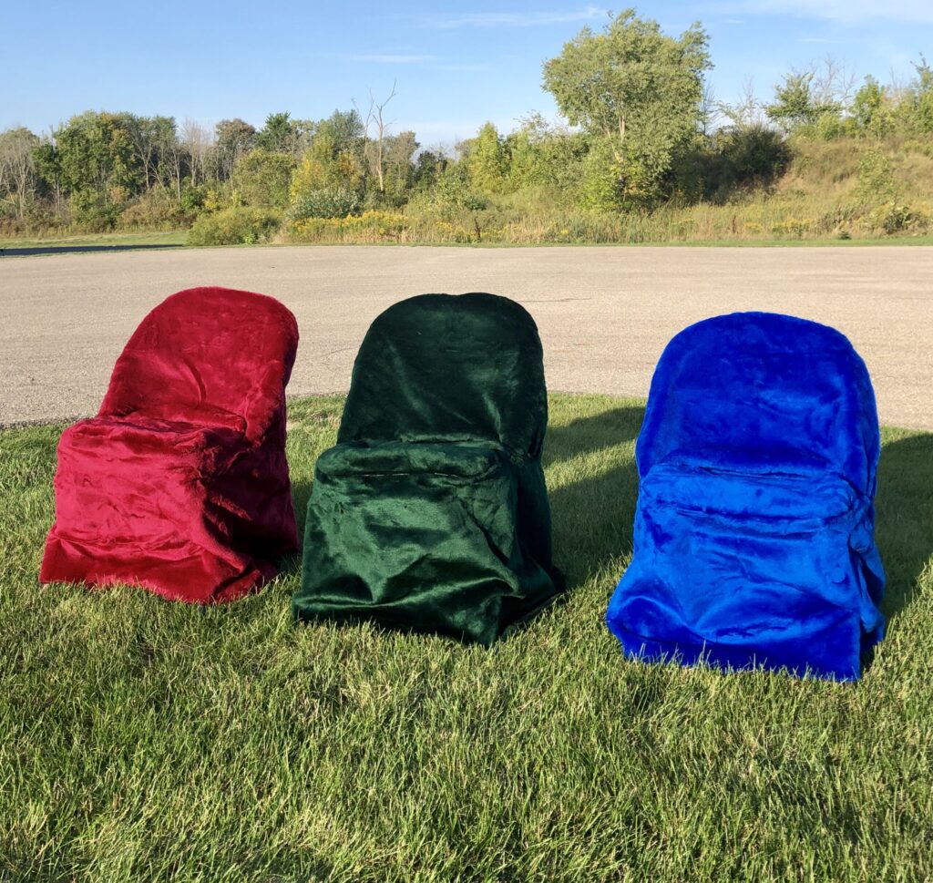 Three chairs covered with fuzzy red, green, and blue fabric sit on a grassy field with trees and a road in the background, subtly blending into nature's tableau like top 10 cemetery products designed for tranquility.