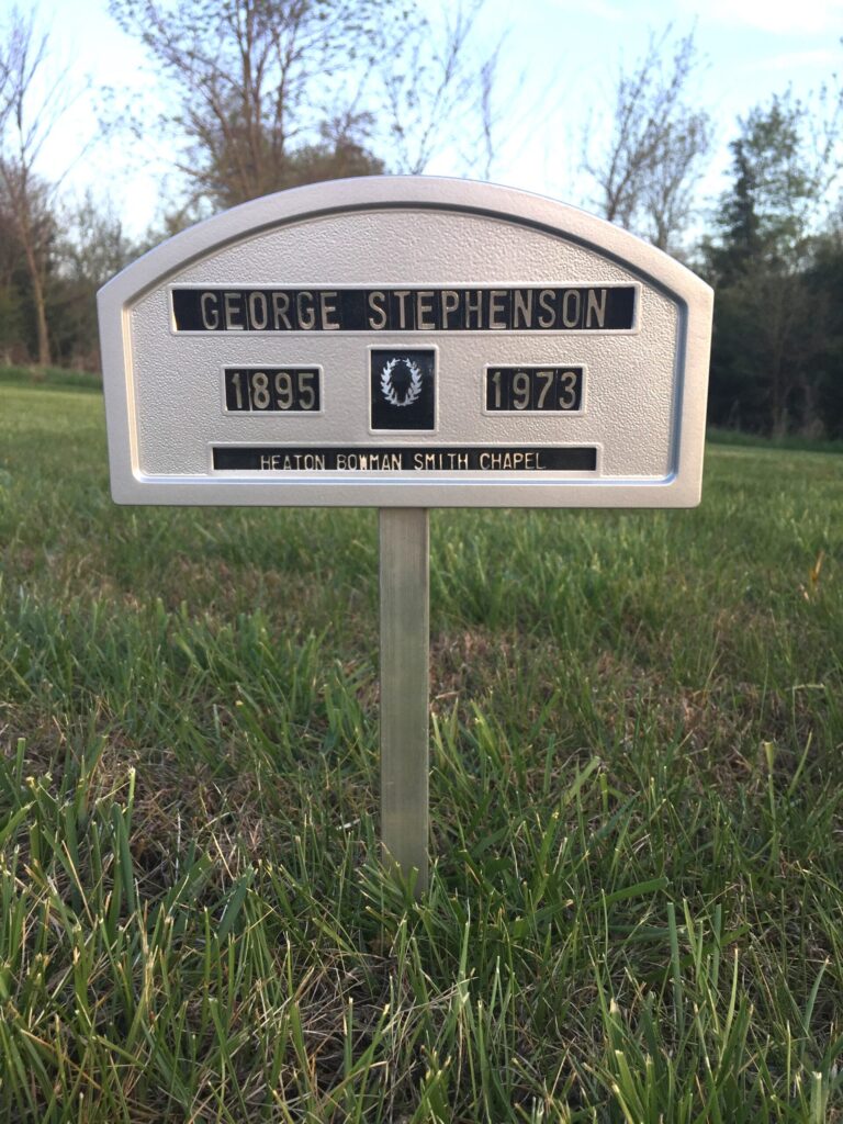 A metal grave marker for George Stephenson, showing the years 1895 and 1973, located on a grassy lawn with trees in the background. It is inscribed with "Heaton Bowman Smith Chapel." This timeless memorial ranks among the top 10 cemetery products for its enduring quality and design.