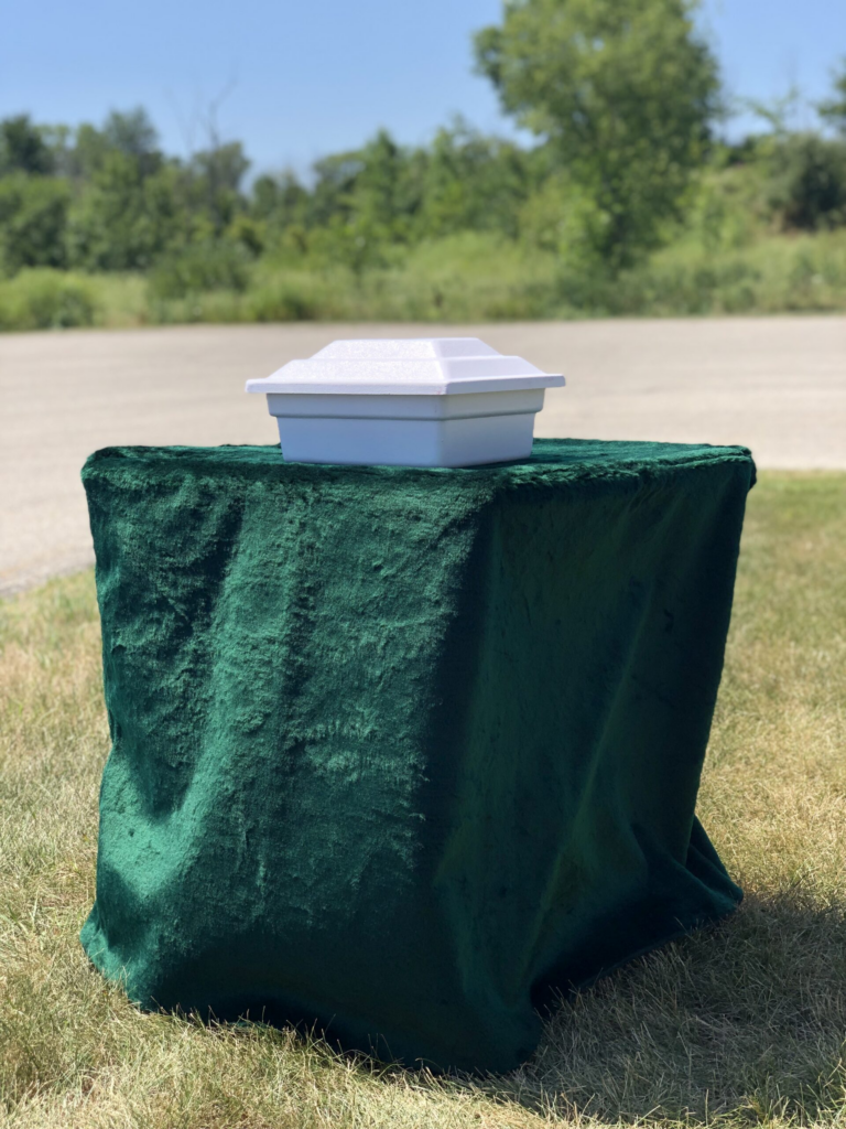 A white takeout container sits on a green cloth-covered table outdoors, with trees and a road in the background, near where old cremation stands once lined the path.