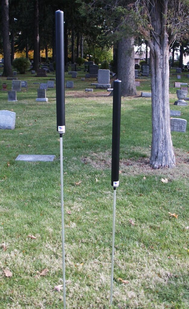 Two black rods, part of our bestselling cemetery products, are mounted on metal poles in a grassy setting with headstones and trees in the background.