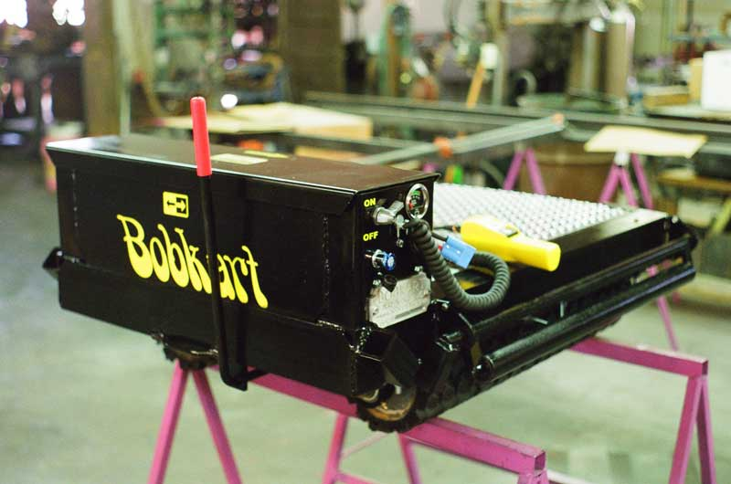 A black machine labeled "Bobkart" with a control panel and red lever sits on a pink stand in a workshop.