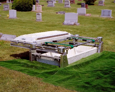 A closed casket is positioned above an open grave with a burial lowering device in a cemetery surrounded by headstones and grass.