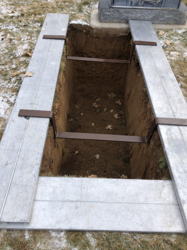 A freshly dug grave with a rectangular hole in the ground, bordered by wooden planks and metal supports, in a cemetery with sparse snow on the grass.