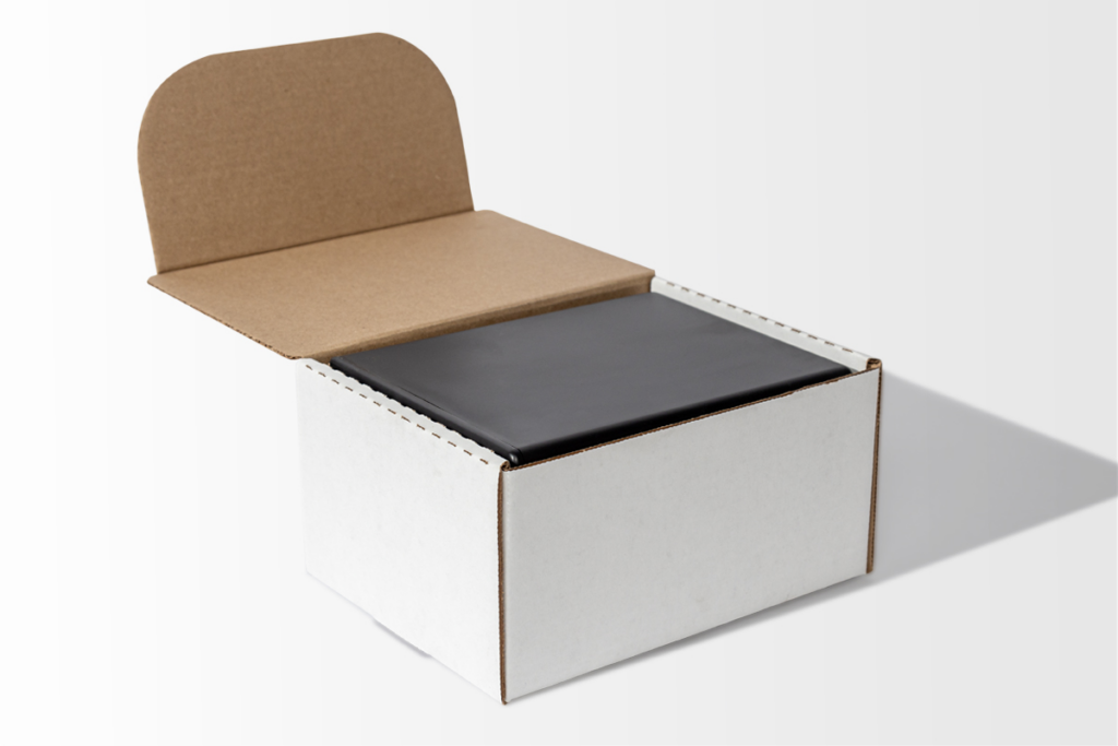 A white cardboard box with an open lid reveals a black rectangular item inside, often used as part of cremation supplies, set against a plain light background.