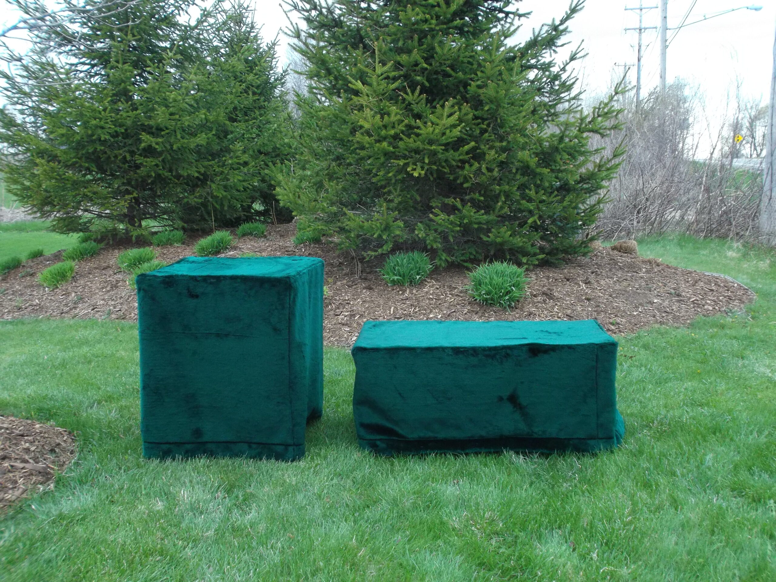 Two green rectangular objects, possibly used as cremation supplies, are covered with fabric and placed on a grassy lawn near some evergreen trees and shrubs in an outdoor setting.