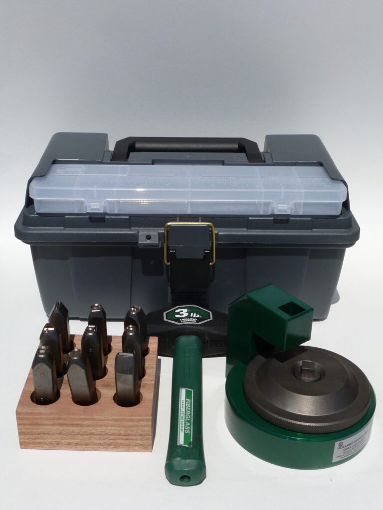 A gray toolbox with a clear lid sits behind a set of metal stamps in a wooden block, cremation supplies, a green handle, and a green metal device on a white surface.