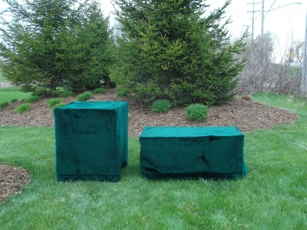 Two rectangular objects covered with green fabric sit on a grassy area in front of pine trees and shrubs in an outdoor setting.
