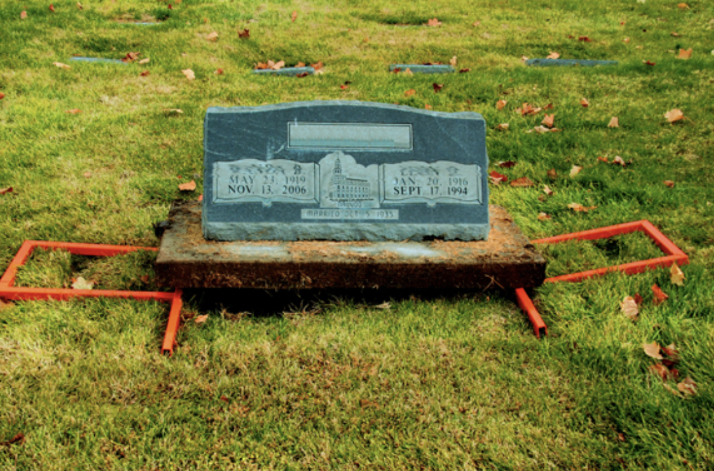 A granite headstone with engraved names and dates stands on a rectangular plot of grass, surrounded by red metal frames.
