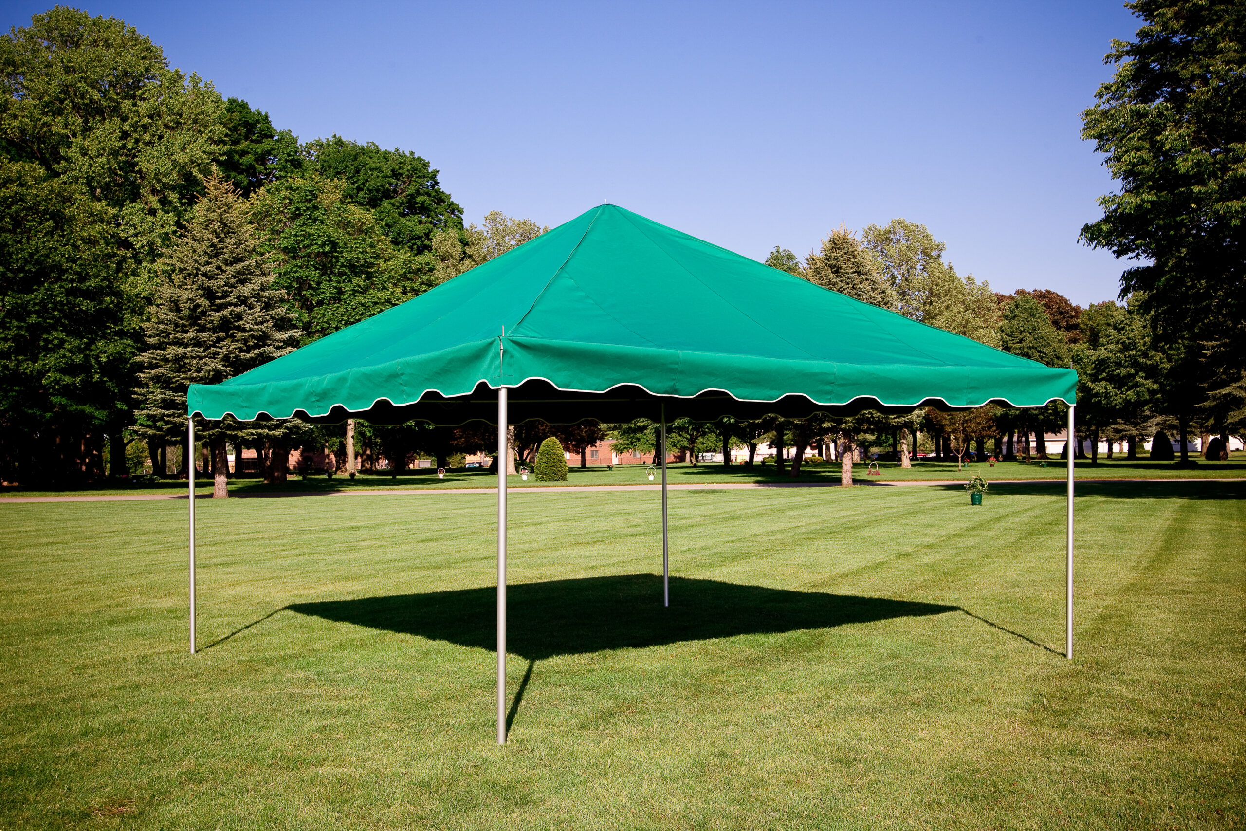 A green canopy tent with white metal legs stands on a grassy field in a park, resembling a graveside setup, surrounded by trees under a clear blue sky.