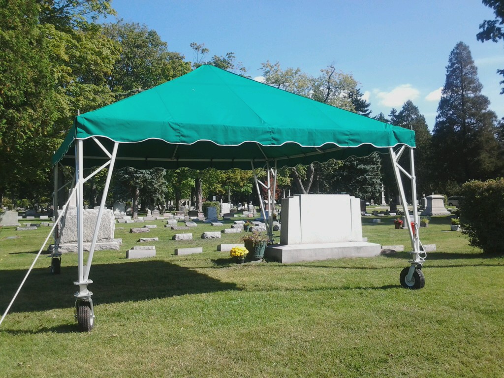 A green canopy tent on wheels is set up in a cemetery, surrounded by gravestones and trees on a sunny day, showing how to streamline graveside setup for ease and efficiency.