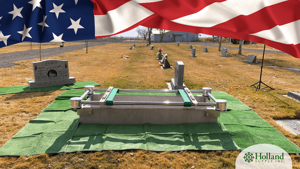 A metal burial vault lowering device positioned over a grave in a cemetery, featuring cemetery equipment with an American flag draped across the top and the Holland Supply Inc. logo in the corner.